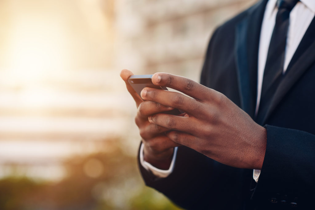 Cropped close up of the hands of an African businessman holding a mobile phone, wiht blurred city building in the background and soft sunflare